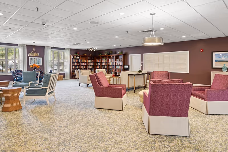 A spacious senior living facility common area with multiple seating arrangements including purple and beige armchairs, blue chairs, and small tables. The room features a carpeted floor, large windows with white curtains, a bookshelf filled with books, and soft lighting from ceiling fixtures. The walls are painted a warm brown color with framed artwork and decorative items.