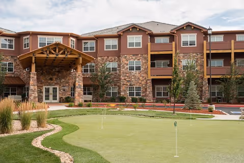 Exterior view of a three-story senior living facility building with stone and wood accents, a covered entrance, and a small putting green with flags in the foreground.