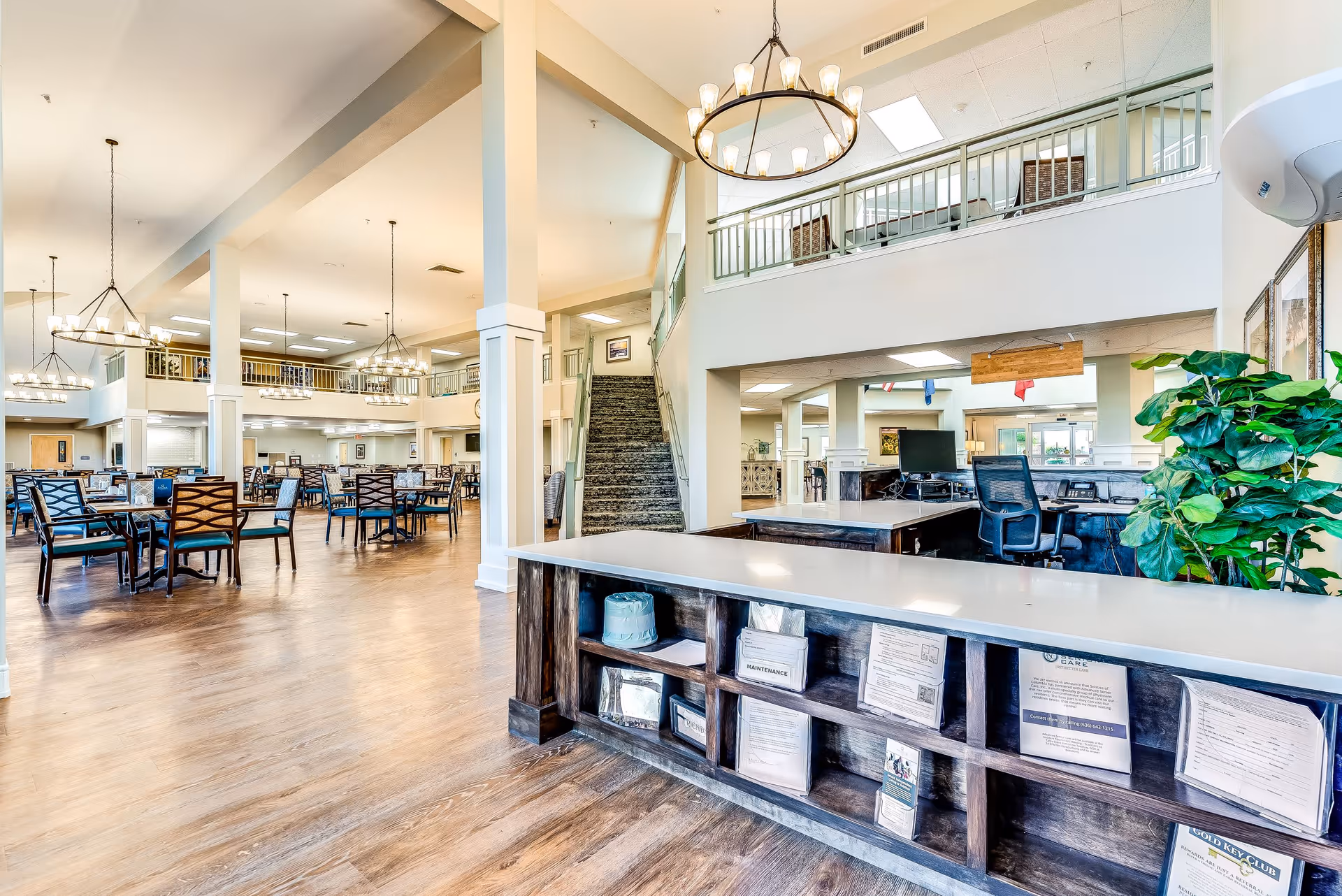 Spacious senior living facility interior featuring a large dining area with multiple tables and chairs, high ceilings with hanging chandeliers, a staircase leading to an upper level, and a reception desk with informational brochures and a potted plant.