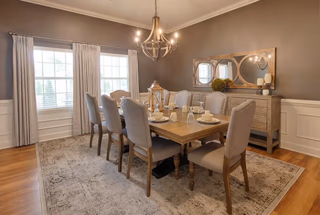 A well-lit dining room with a wooden dining table set for six, surrounded by upholstered chairs. The room features large windows with light curtains, a chandelier hanging above the table, a decorative rug underneath, and a sideboard with a large mirror and decorative items on top.