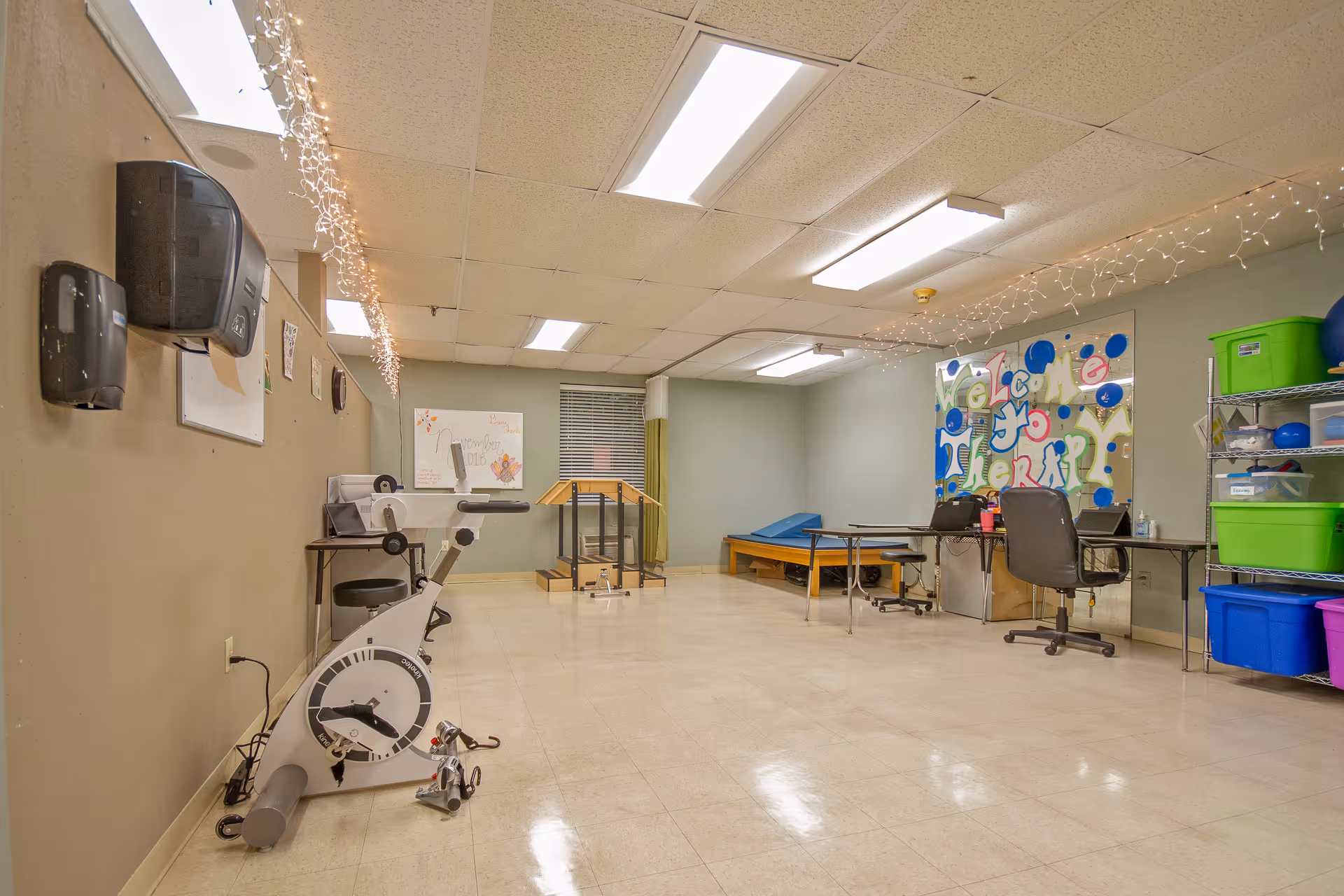 Empty therapy room with an exercise bike, treatment tables, desks, storage bins and a colorful 'Welcome to Therapy' mural.