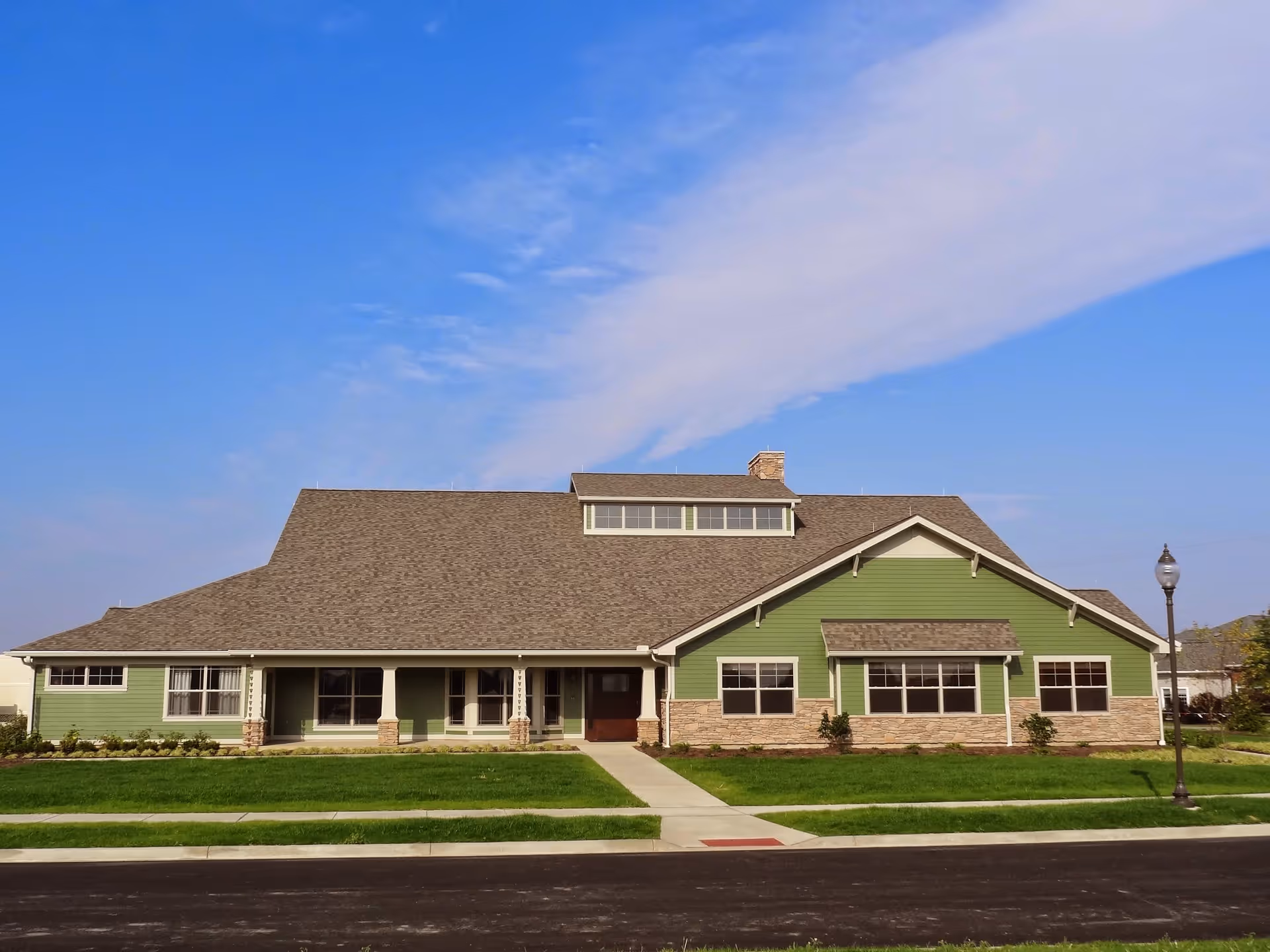 Front exterior view of a single-story building with green siding and stone accents, a large sloped roof with a dormer window, a sidewalk leading to the entrance, and a streetlamp on the right side under a blue sky with some clouds.