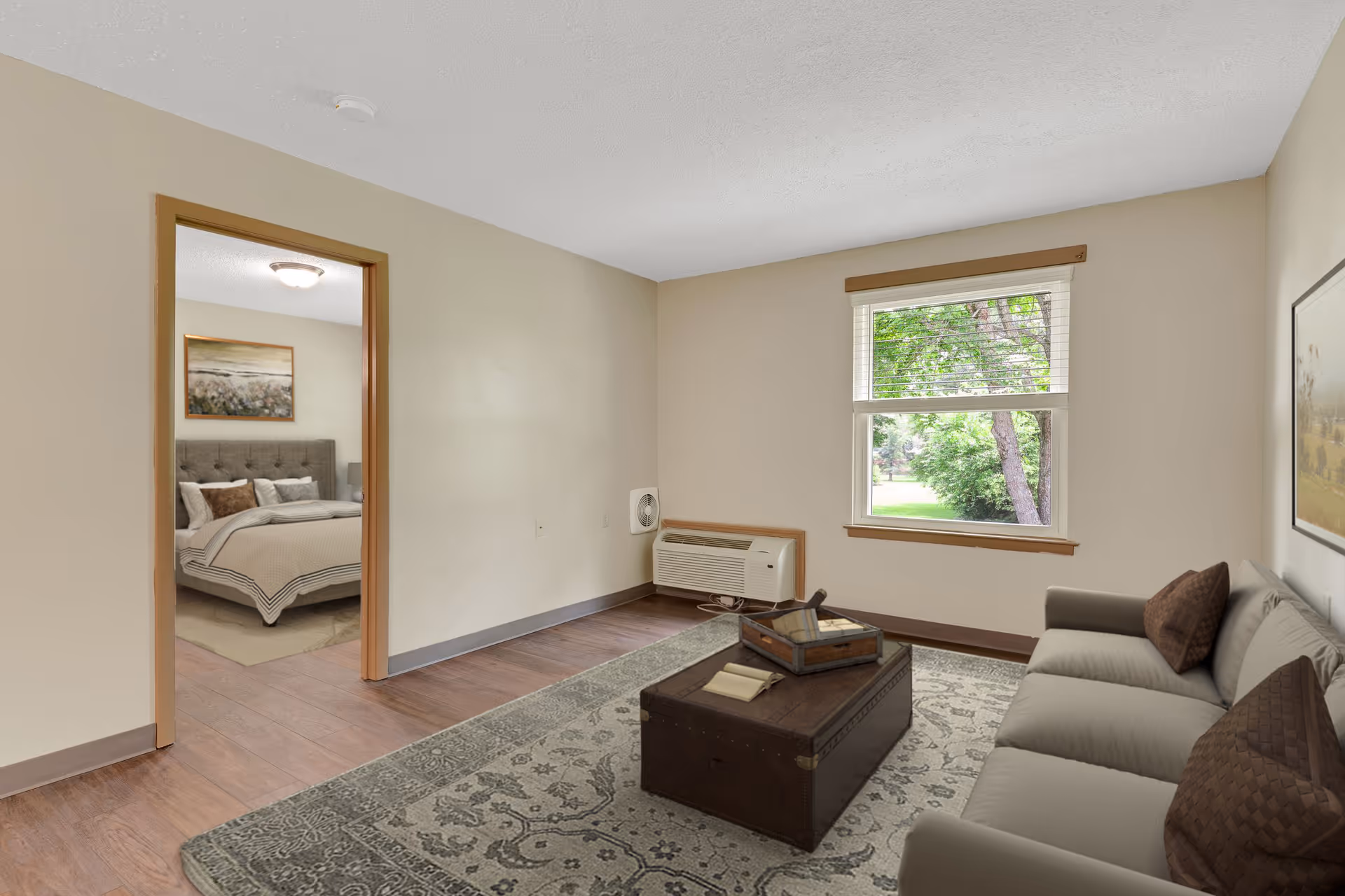 A cozy living room with a gray sofa adorned with brown pillows, a wooden chest used as a coffee table on a patterned rug, and a window showing green trees outside. Through an open doorway, a bedroom with a neatly made bed and a framed picture above the headboard is visible.