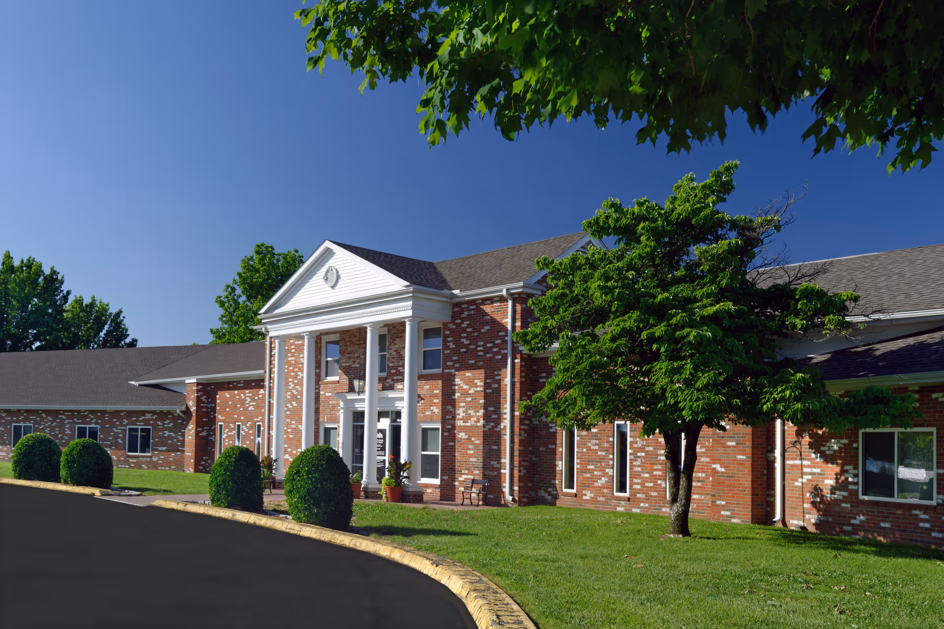 Exterior view of Community Springs Healthcare Facility showing a large brick building with white columns at the entrance, surrounded by green grass, bushes, and trees under a clear blue sky.