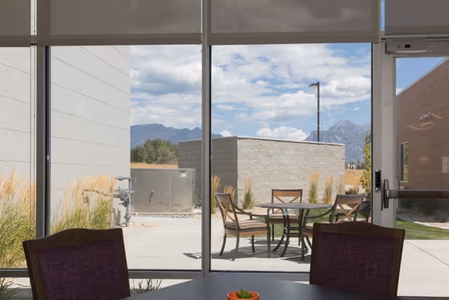 View through large glass windows showing an outdoor patio area with a round table and four chairs. The patio is surrounded by concrete walls and landscaping with tall grasses. Mountains and a partly cloudy sky are visible in the background.