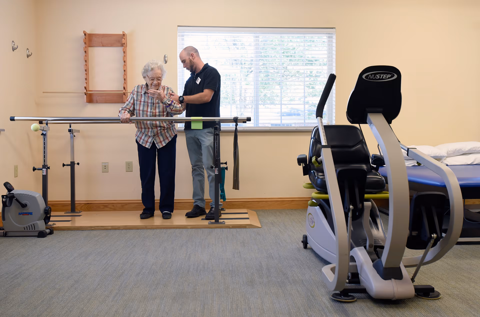 An elderly woman is assisted by a male caregiver or physical therapist as she uses parallel bars for walking exercises in a rehabilitation or therapy room. The room has exercise equipment including a stationary bike and a NuStep machine, with a window in the background letting in natural light.