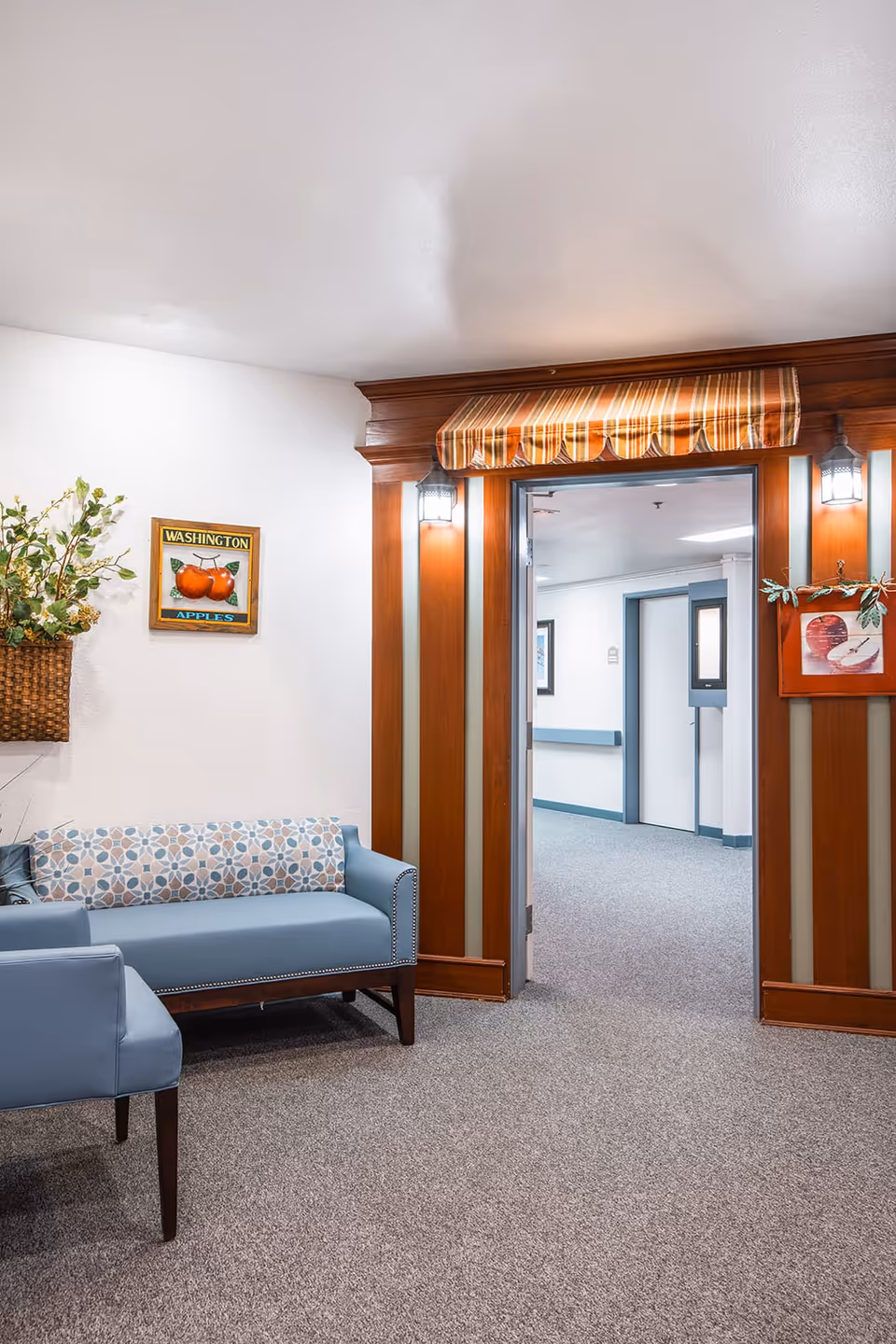 A cozy interior seating area with a blue upholstered loveseat and chair on a carpeted floor. The wall features a basket with greenery and a framed picture of Washington apples. A wooden doorway with striped fabric awning and two lantern-style lights leads to a hallway with white walls and blue trim.