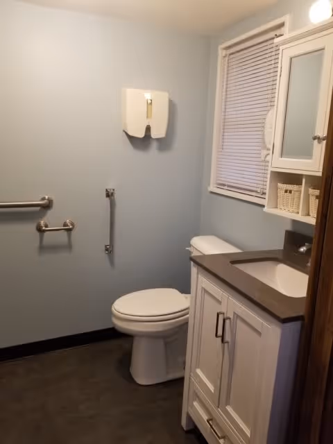 A clean bathroom with a white toilet, a white vanity with a gray countertop and built-in sink, a wall-mounted cabinet with baskets, a window with closed blinds, and grab bars on the wall for accessibility.