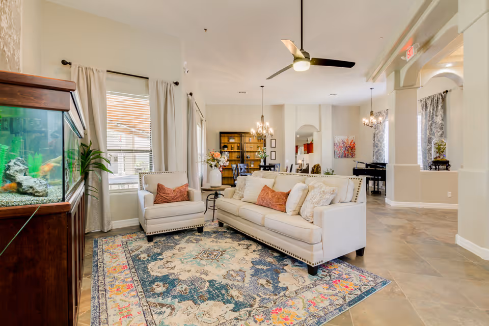 Bright and spacious living room area with a beige sofa and armchair adorned with cushions, a colorful patterned rug, a large fish tank on a wooden cabinet, ceiling fan, chandeliers, and large windows with curtains letting in natural light. In the background, there is a dining area with a wooden bookshelf and a grand piano near arched windows.