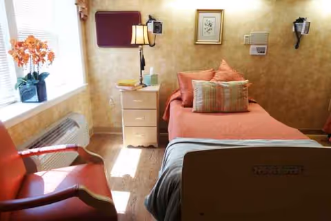 A cozy hospital bedroom with a bed, nightstand, lamp, red chair, and sunlit window with flowers.