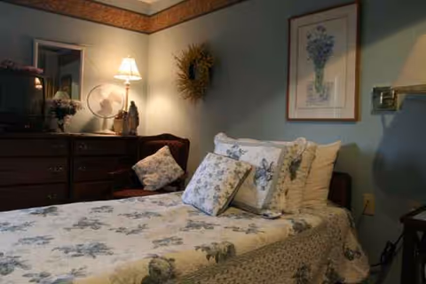 A cozy bedroom with a floral quilt, pillows, dresser, lamp, and wall art.