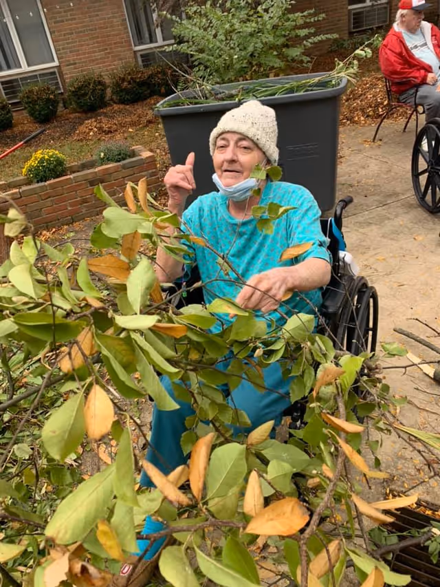 An elderly woman in a wheelchair wearing a knitted hat and teal clothing is outdoors, holding and interacting with leafy branches. Another elderly person in a red jacket and gray pants is seated in a wheelchair in the background near a brick building with windows and some plants.