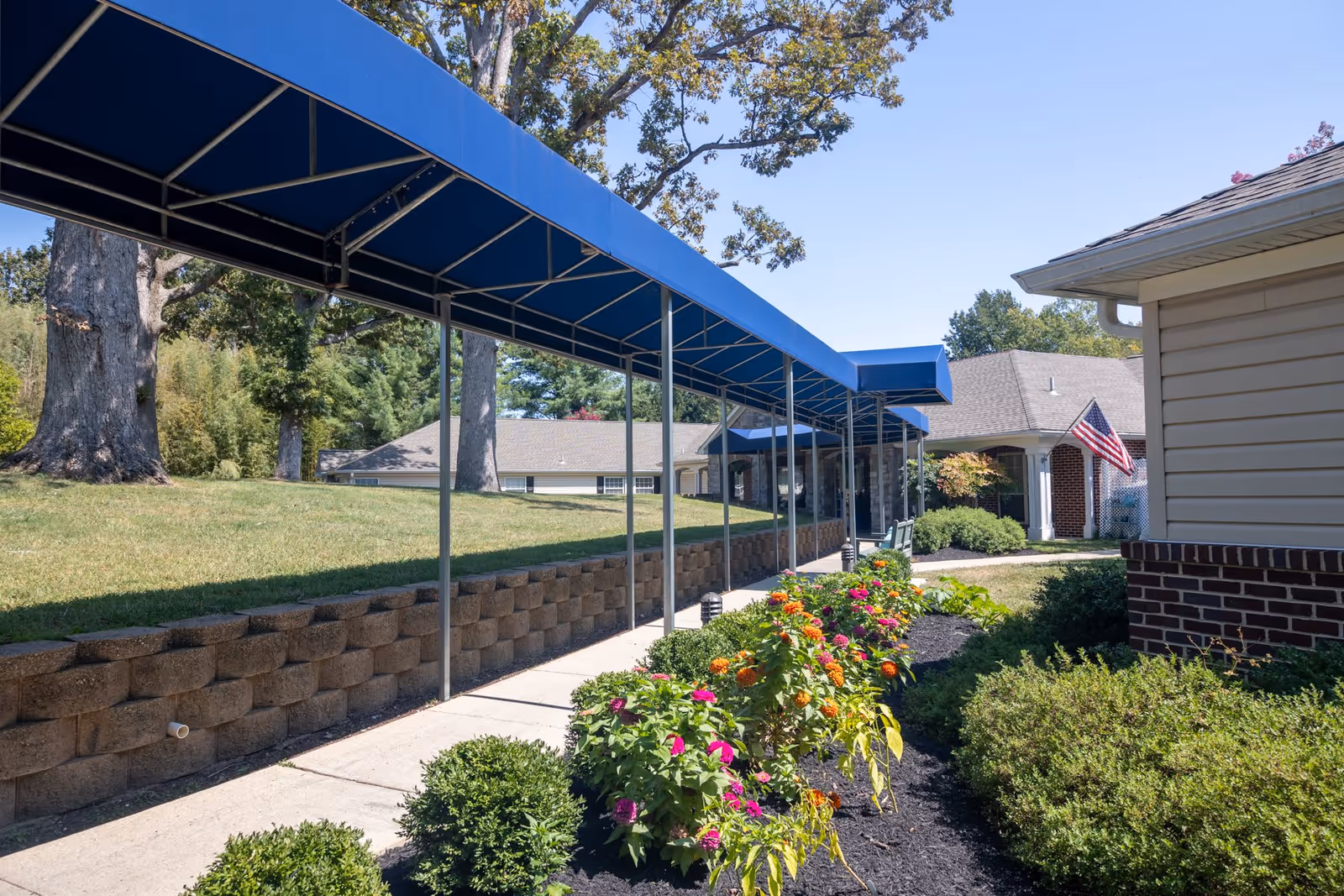 Outdoor walkway at Charter Senior Living of Annapolis with a blue canopy covering a concrete path. The path is bordered by a flower bed with colorful flowers and green shrubs on one side and a retaining wall with grass and large trees on the other. Residential-style buildings with beige siding and brick accents are visible in the background, along with an American flag near the entrance.