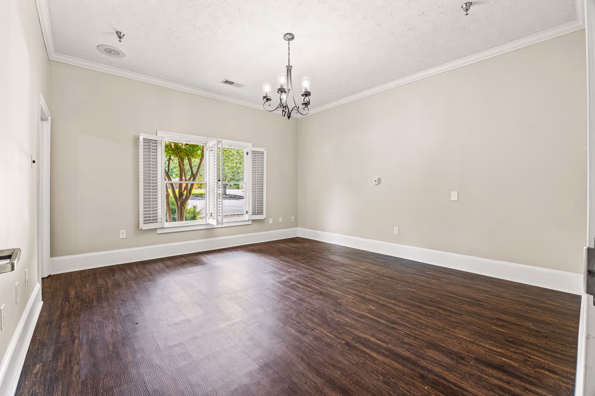 Empty room with dark wood floors, neutral beige walls, a hanging chandelier, and a window with white shutters showing trees outside.