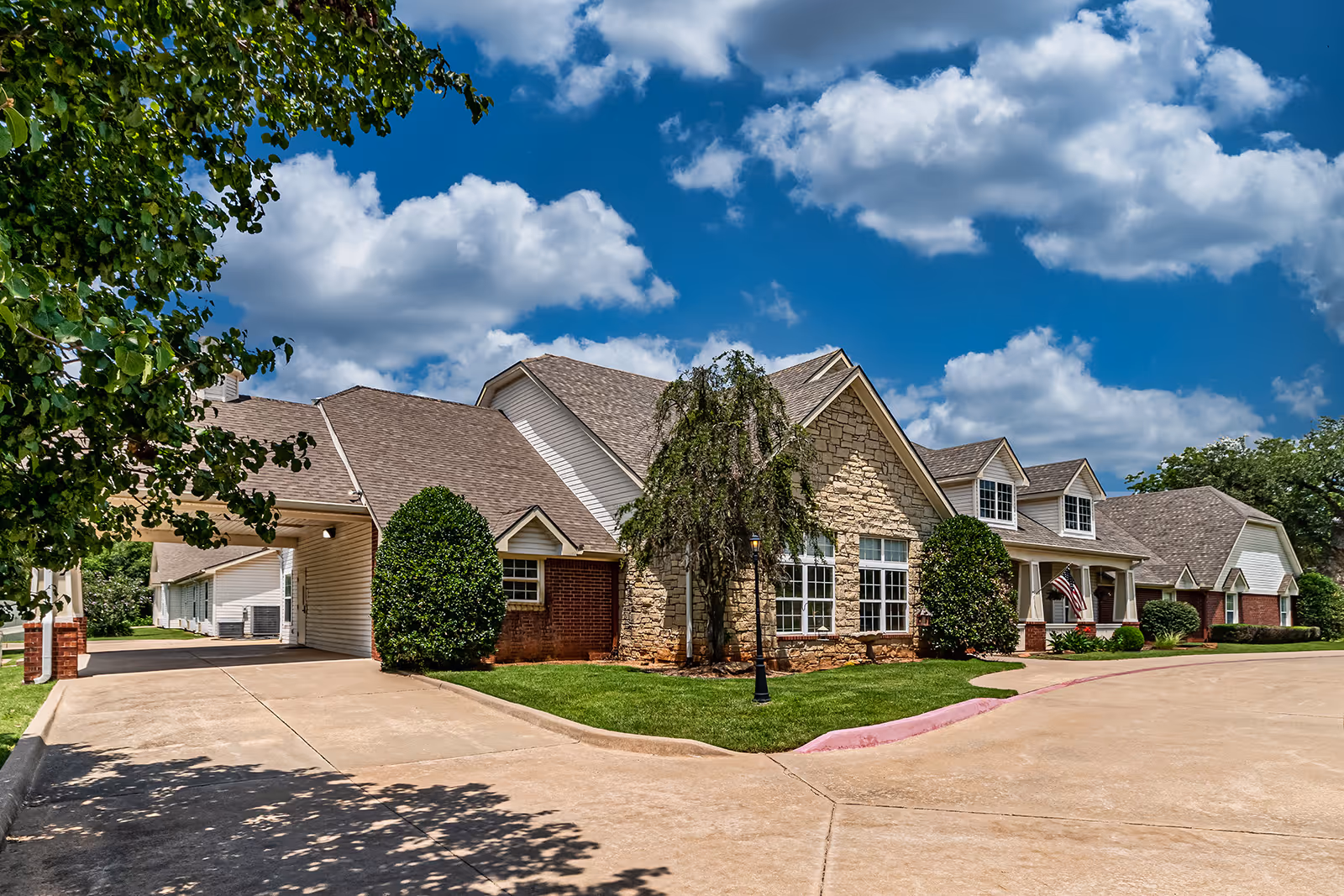 Exterior view of Ashbrook Village facility showing a large building with a mix of stone, brick, and siding on the walls, multiple gabled roofs, and several windows. The building is surrounded by neatly trimmed bushes and a green lawn. A driveway curves in front of the building under a partly cloudy blue sky.