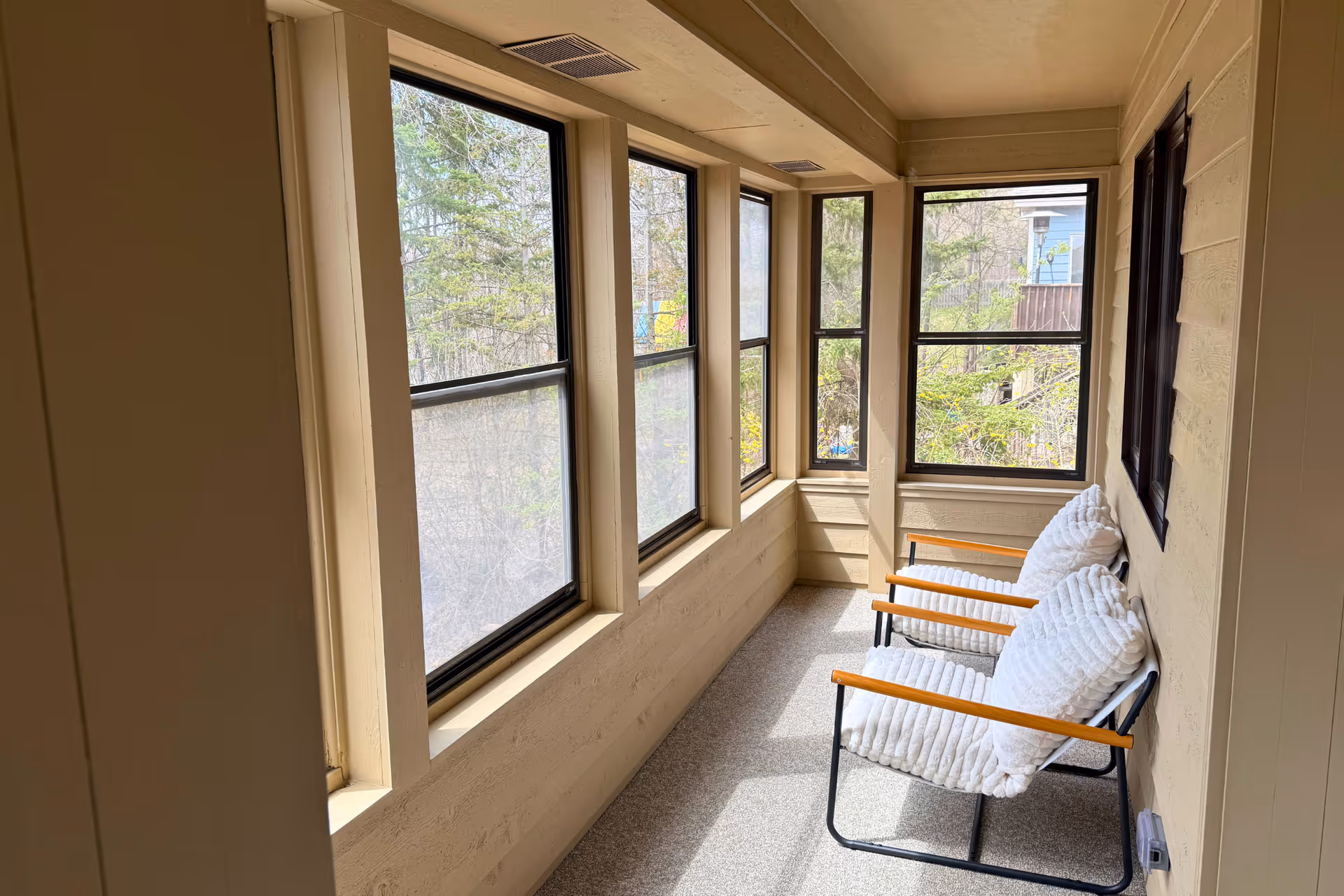 A narrow sunroom or enclosed porch area with large windows on two sides, allowing natural light to fill the space. Two cushioned chairs with wooden armrests are placed against the wall, facing the windows. Outside, trees and parts of neighboring buildings are visible.