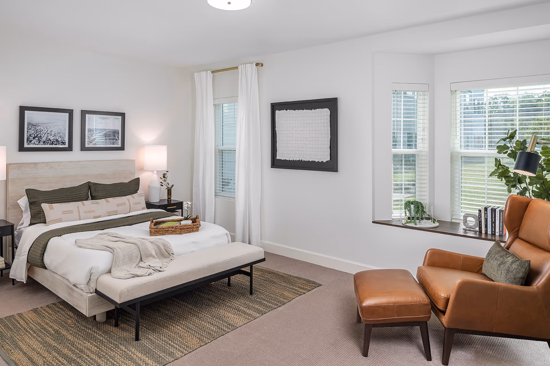 Bright modern bedroom with a made bed, bench at the foot, leather armchair and a bay window with plants.
