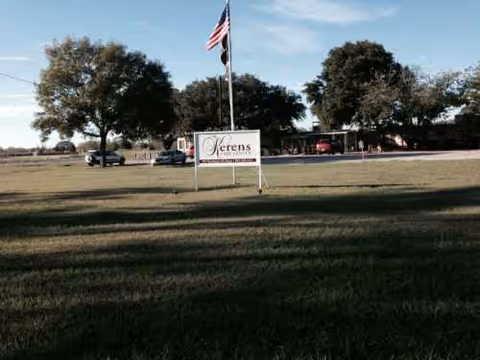 A grassy exterior scene with a Kerens Care Center sign, flagpole, trees, and a distant building.