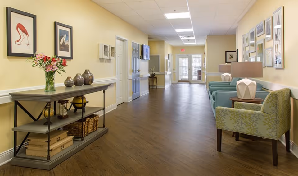 A well-lit hallway in a senior living facility with wooden flooring, light yellow walls, and a white ceiling. On the left side, there is a console table with decorative vases, flowers, and baskets underneath. On the right side, there are several upholstered chairs and side tables with lamps. Framed artwork is hung on the walls, and double glass doors are visible at the end of the hallway.