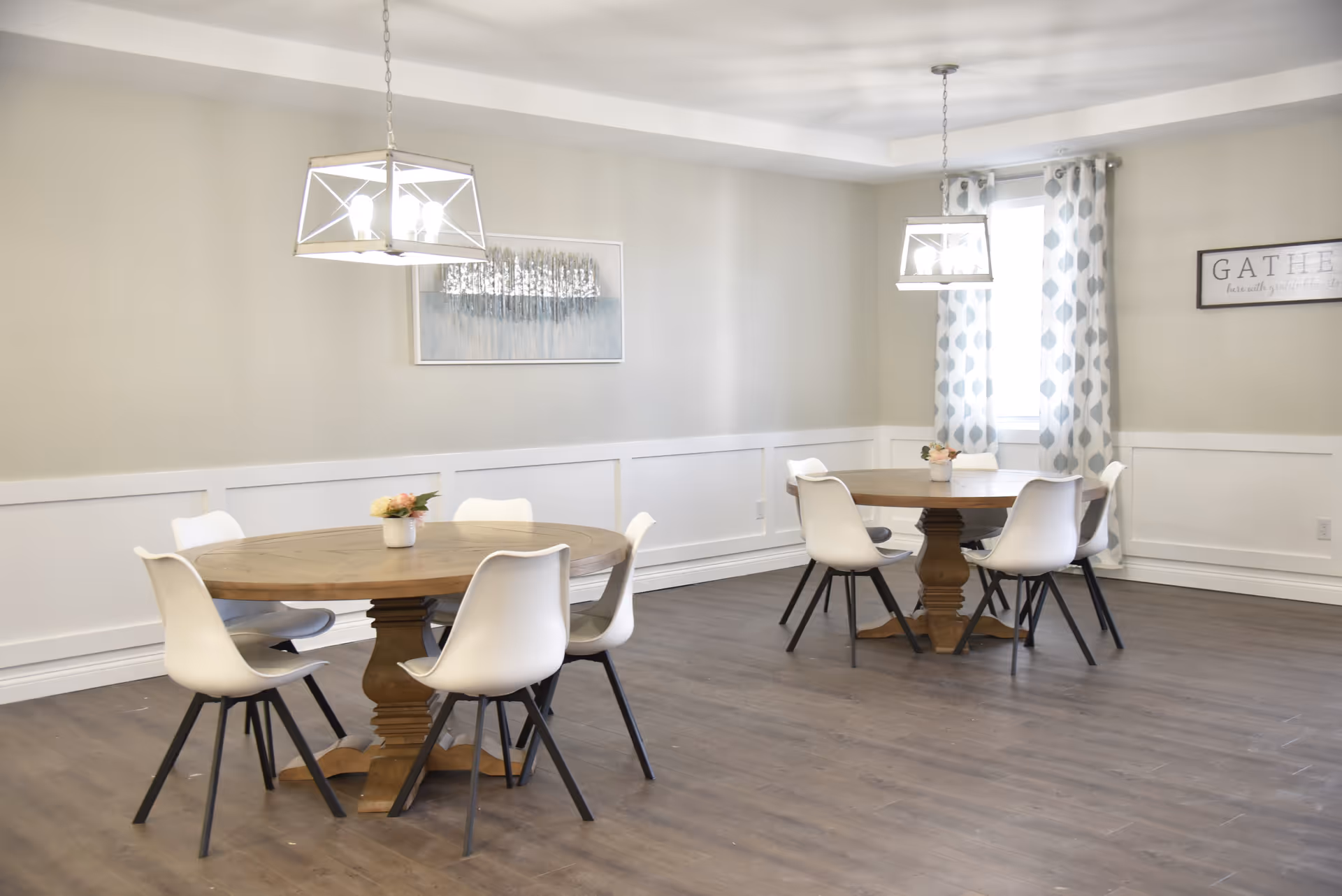 A bright dining room with two round wooden tables, each surrounded by six white chairs with black legs. Each table has a small flower arrangement in the center. The room has light-colored walls with white wainscoting, two modern pendant lights hanging from the ceiling, a window with patterned curtains, and framed wall art including a sign that says 'GATHER'.
