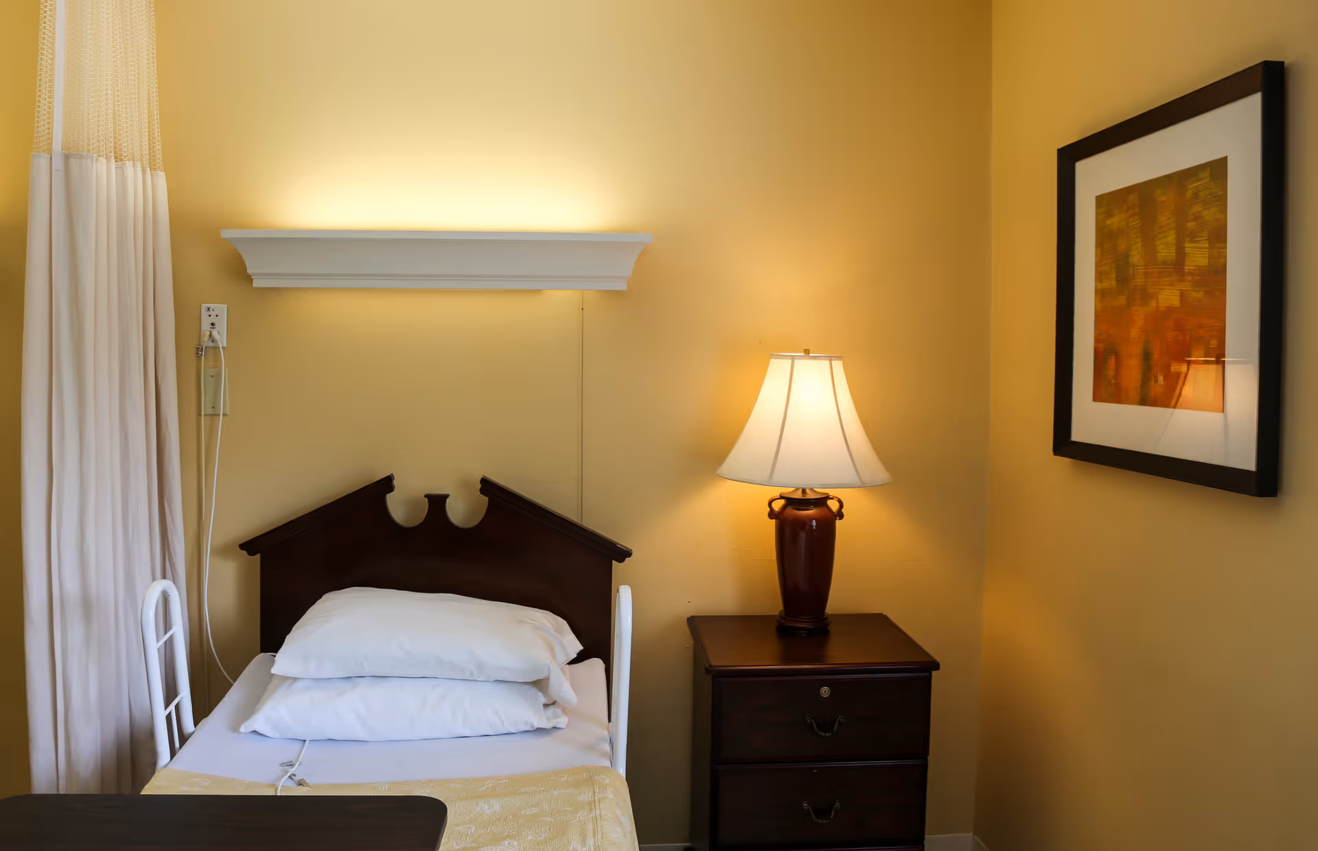 A single bed with white pillows and a yellow blanket in a room with yellow walls. Next to the bed is a dark wooden nightstand with a lamp on it. A framed abstract painting hangs on the wall above the nightstand. A white curtain is partially visible on the left side of the image.