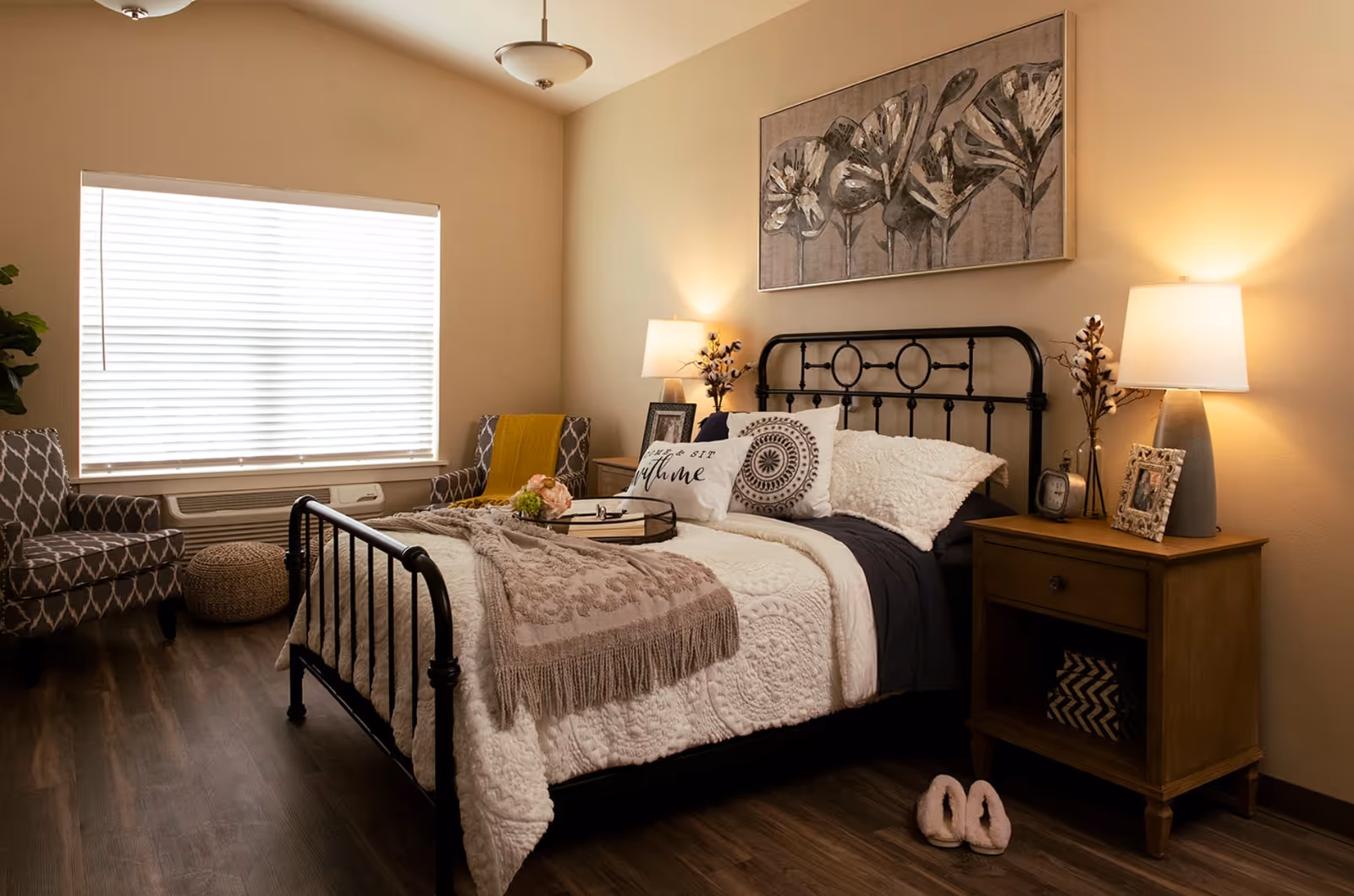 Cozy bedroom featuring a black metal bed with decorative pillows and throw, a wooden nightstand with a lamp, and an armchair by a window.