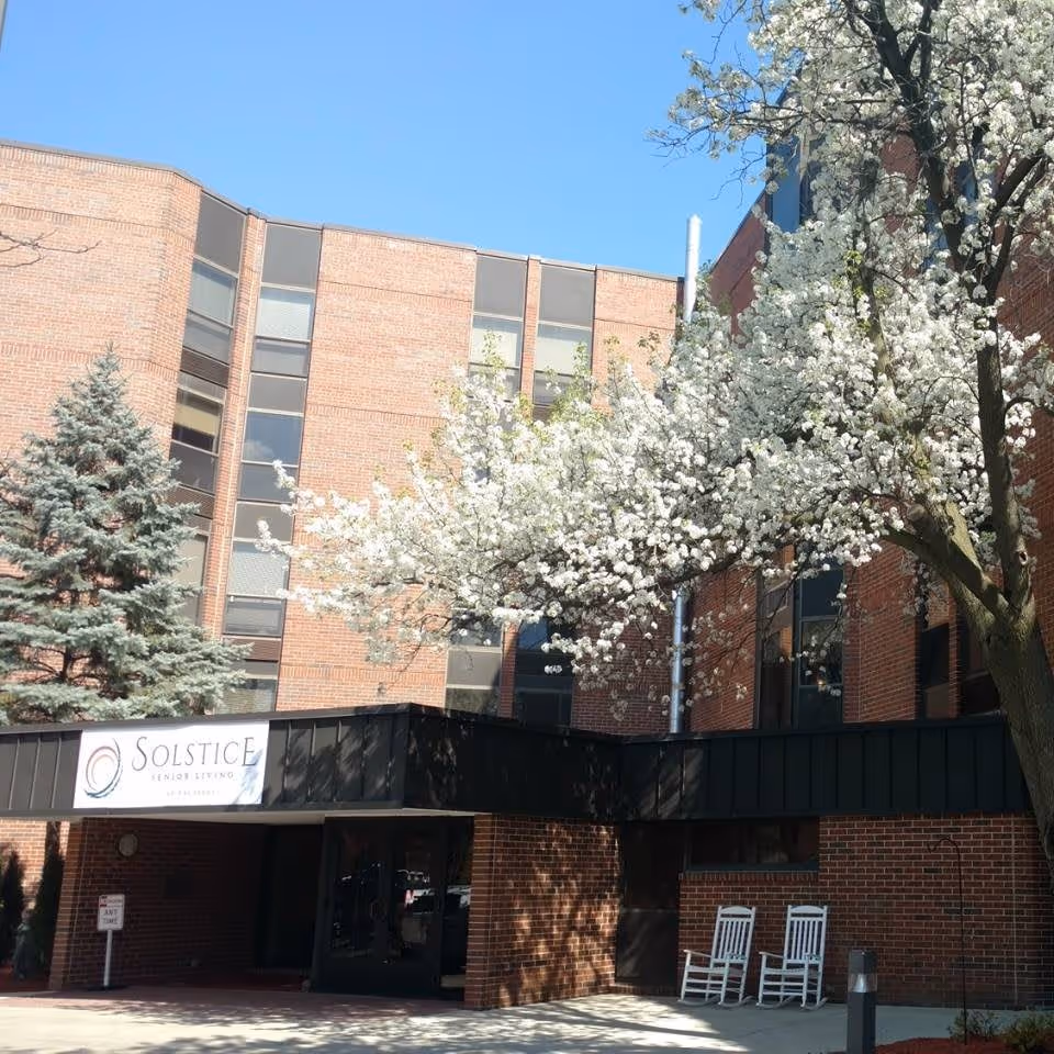 Exterior view of Solstice Senior Living at Palatine building with a brick facade, large windows, a black awning over the entrance, two white rocking chairs on the porch, and a tree with white blossoms in the foreground under a clear blue sky.