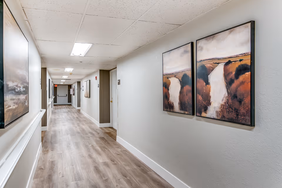 A clean, well-lit hallway in a senior living facility with light wood flooring, white baseboards, and beige walls. The ceiling has recessed fluorescent lighting. On the right wall, there are two framed landscape paintings depicting a river flowing through autumn-colored trees. On the left wall, there are additional framed artworks. Several doors line the hallway, and an exit sign is visible at the far end.