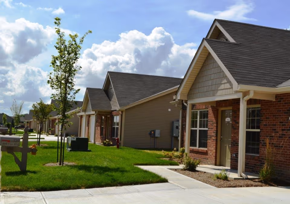 Row of single-story residential buildings with brick and siding exteriors, small front lawns with young trees, and a clear blue sky with clouds above.