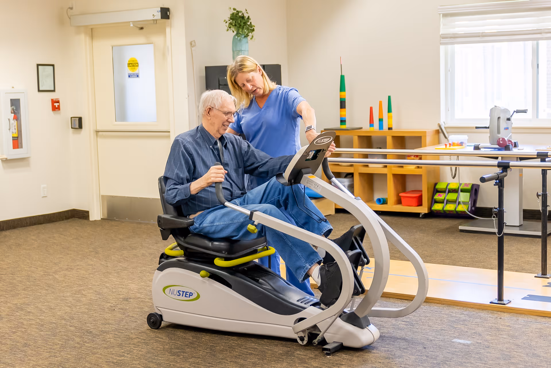 An elderly man is seated on a NuStep exercise machine in a well-lit room, assisted by a female caregiver in blue scrubs who is standing beside him and providing support. The room has carpeted flooring, a window with blinds, and shelves with colorful objects and exercise equipment in the background.