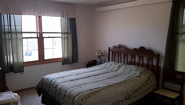 A bedroom with a wooden bed frame and a green patterned bedspread. There are two windows with green curtains on either side of the bed, allowing natural light into the room. A small bedside table with a lamp is visible next to the bed.