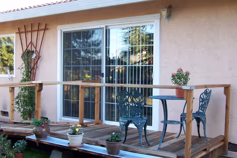 Outdoor deck with railing, small table and chairs, and potted plants in front of glass doors.