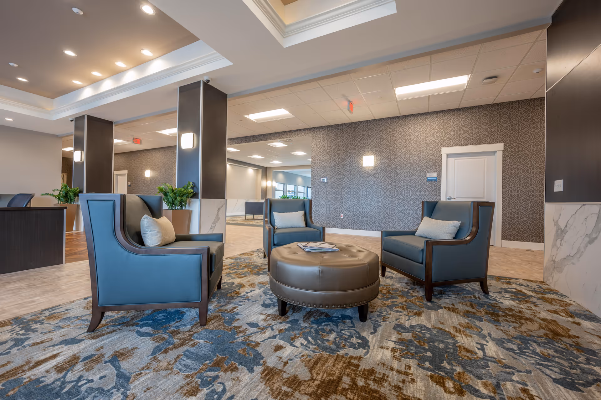A modern and spacious seating area in a senior living facility with three blue armchairs arranged around a round brown ottoman on a patterned carpet. The room features decorative wallpaper, recessed lighting, potted plants, and a ceiling with tray design and multiple light fixtures.