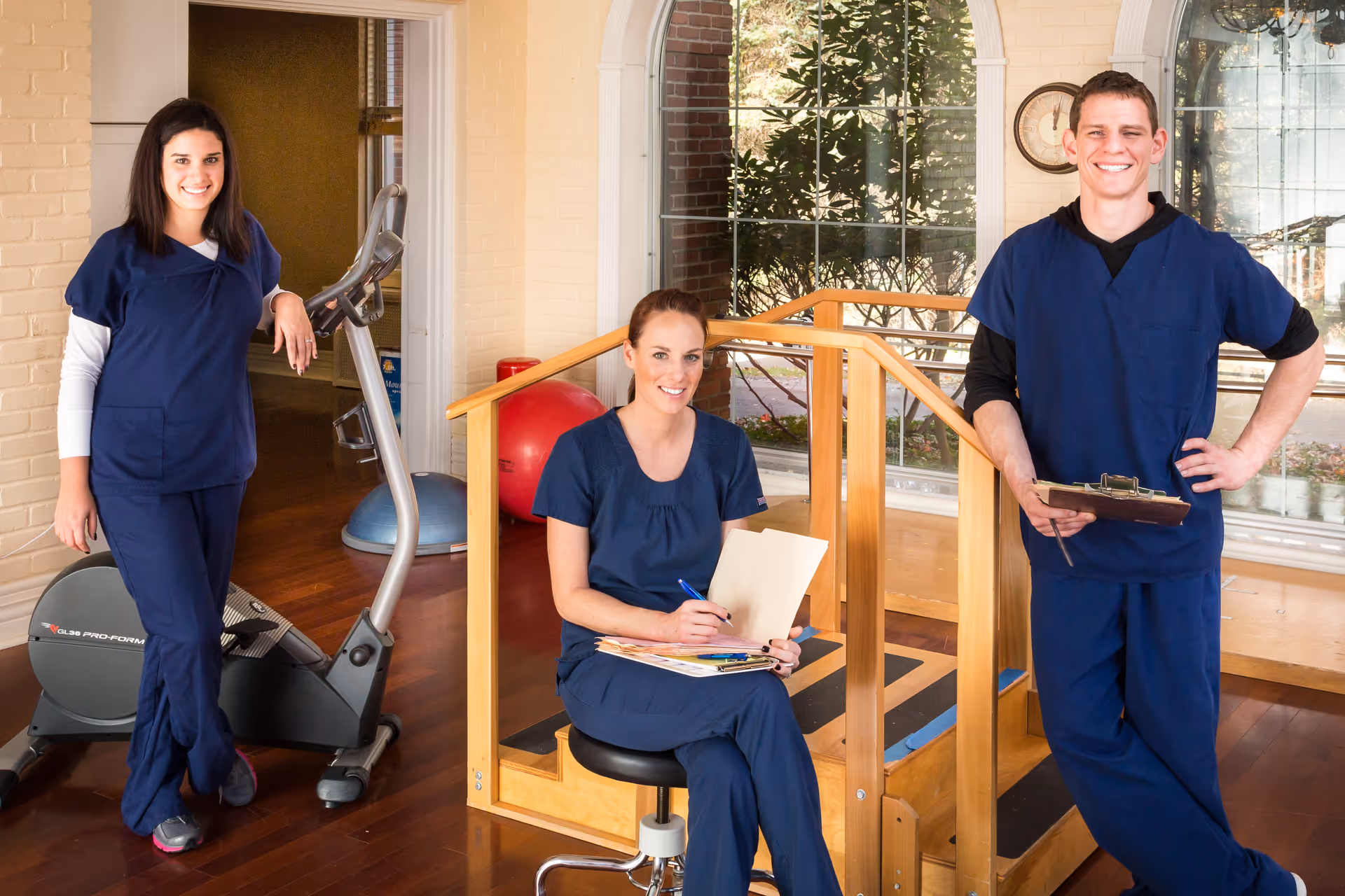 Three healthcare professionals in navy blue scrubs posing in a rehabilitation or therapy room with exercise equipment and wooden handrails. One woman is sitting on a stool holding a clipboard and pen, while the other two stand nearby smiling.