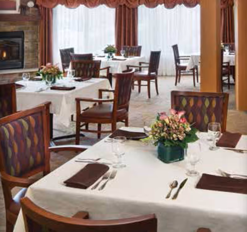 A dining room with multiple tables covered in white tablecloths, each set with brown napkins, silverware, glasses, and floral centerpieces. The room has large windows with sheer white curtains and brown drapes, and a stone fireplace is visible on the left side.