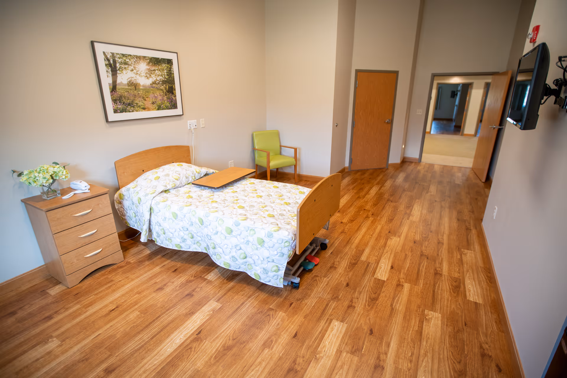 A clean and simple bedroom in a senior living facility with a single bed covered in a patterned bedspread, a wooden nightstand with a telephone and flowers, a green chair, a wall-mounted TV, and a framed picture on the wall. The room has wooden flooring and neutral-colored walls.