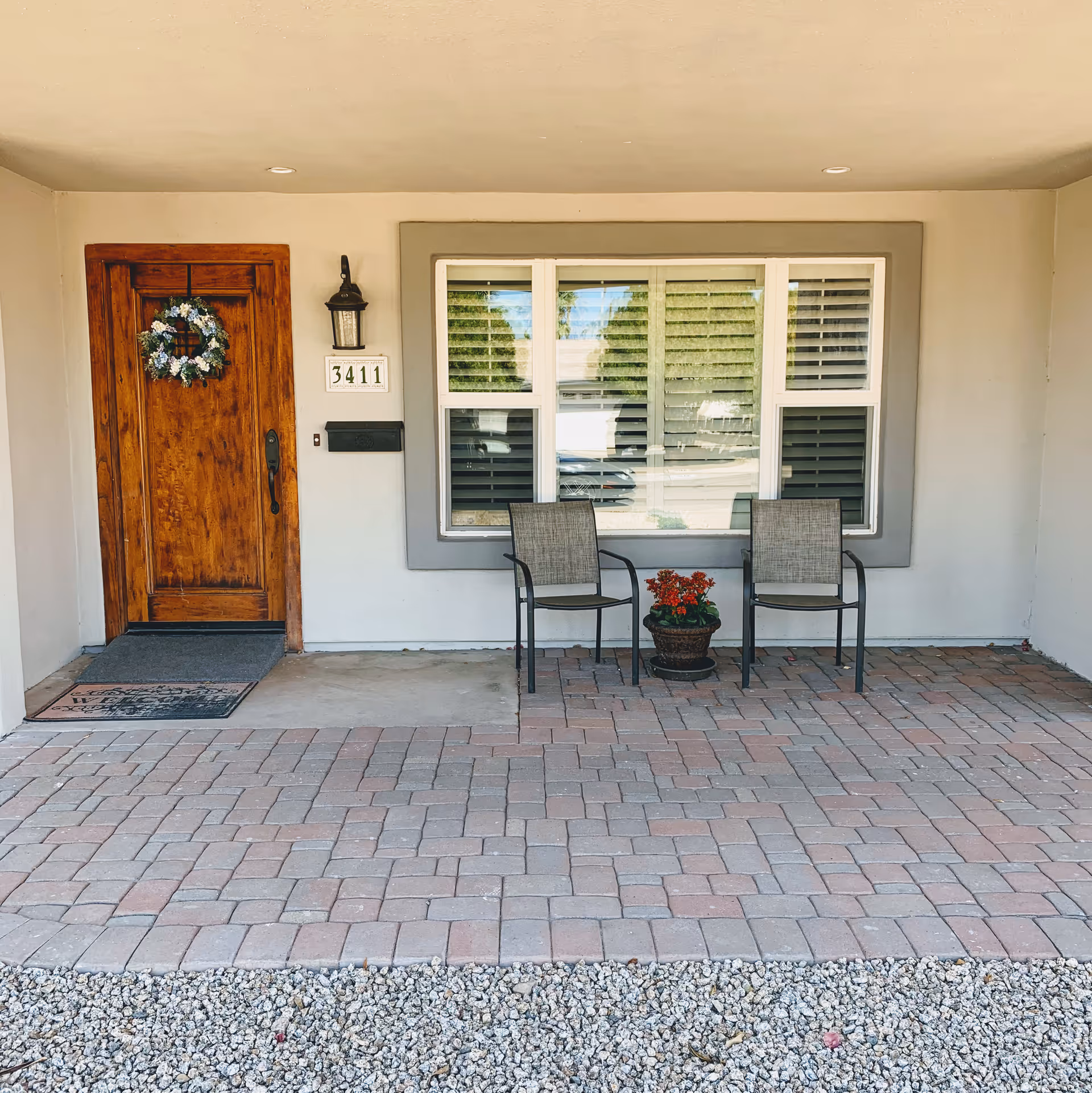 Covered front porch area with a wooden door decorated with a floral wreath, a wall-mounted lantern light, house number 3411, a mailbox, two patio chairs, and a potted plant placed between the chairs. The porch floor is paved with bricks and gravel is visible in the foreground.