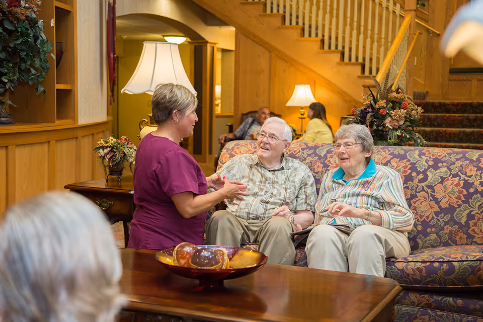A caregiver in a purple uniform is talking to an elderly man and woman seated on a floral-patterned couch in a warmly lit living room with wooden paneling and a staircase in the background. There are table lamps and floral arrangements adding to the cozy atmosphere.