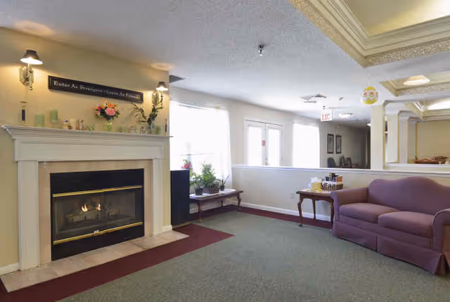A cozy living room area with a lit fireplace on the left, decorated with flowers and small ornaments on the mantel. Above the fireplace is a sign that reads 'Enter As Strangers - Leave As Friends.' To the right, there is a purple upholstered couch and a small wooden side table with items on it. The room has large windows letting in natural light and a ceiling with decorative molding.