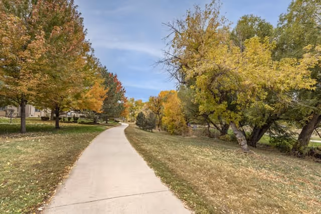 A paved walking path surrounded by grass and trees with autumn foliage in shades of green, yellow, and orange under a partly cloudy sky.
