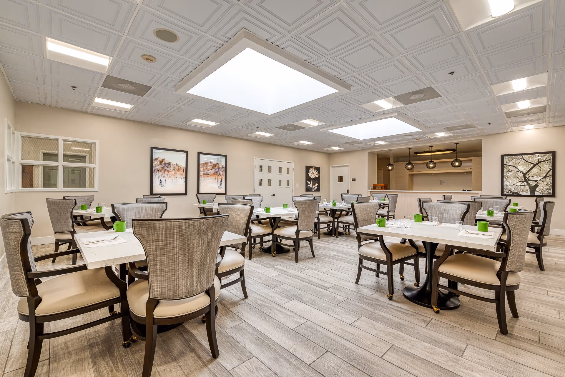 A bright and spacious dining room with multiple tables and cushioned chairs arranged neatly. Each table is set with white napkins, glasses, and small green cups. The room features light-colored wood flooring, a white coffered ceiling with large recessed lights, and framed artwork on the walls. There is a counter area with pendant lights in the background.