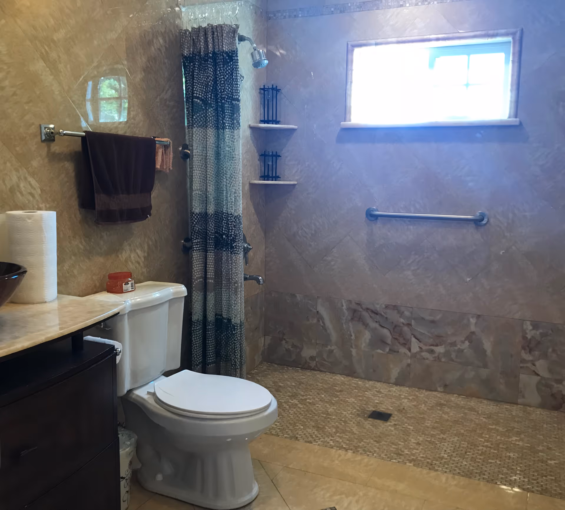 Accessible bathroom with a toilet, countertop sink, patterned shower curtain, wall grab bar, and a small high window.
