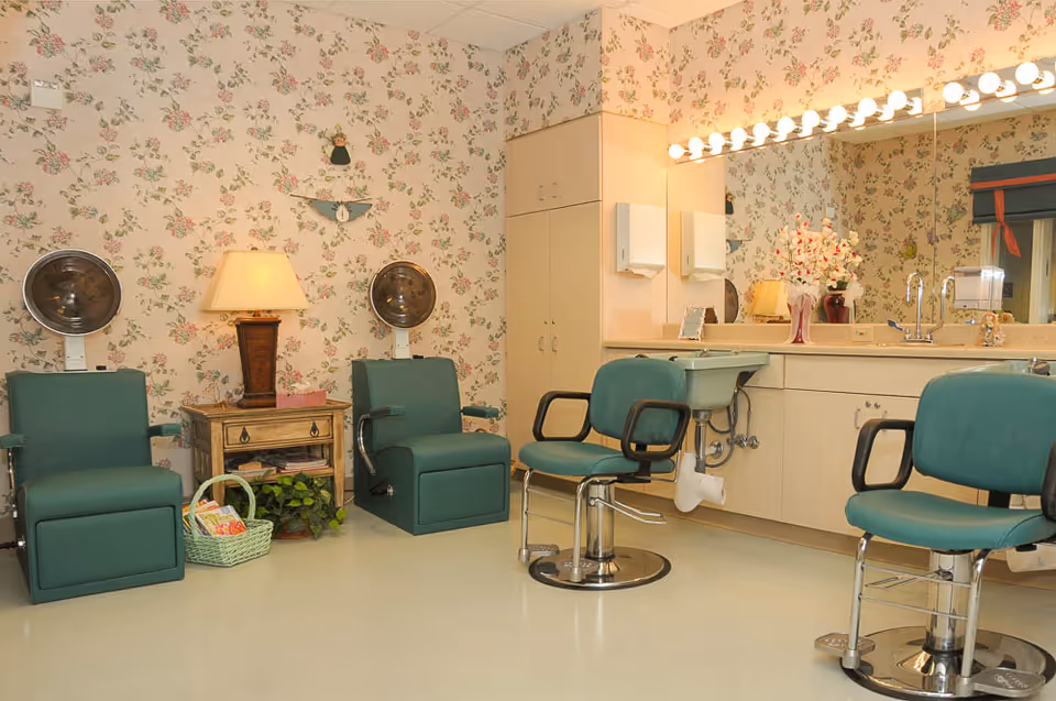 A salon area in a healthcare facility with floral wallpaper, two green hair drying chairs with hooded dryers, two green salon chairs in front of a counter with a large mirror and bright vanity lights. There is a wooden side table with a lamp and a basket with magazines, and a vase with flowers on the counter.