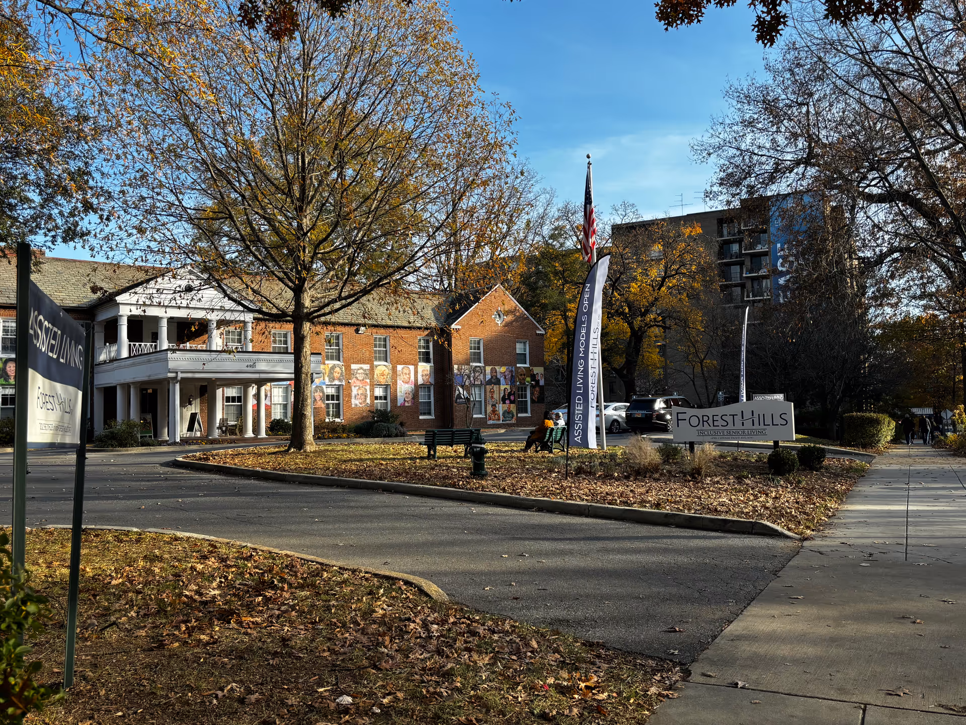 Exterior view of Forest Hills senior living facility with a brick building, trees with autumn leaves, a curved driveway, and signs indicating assisted living and inclusive senior living. Two people are sitting on a bench near the entrance.