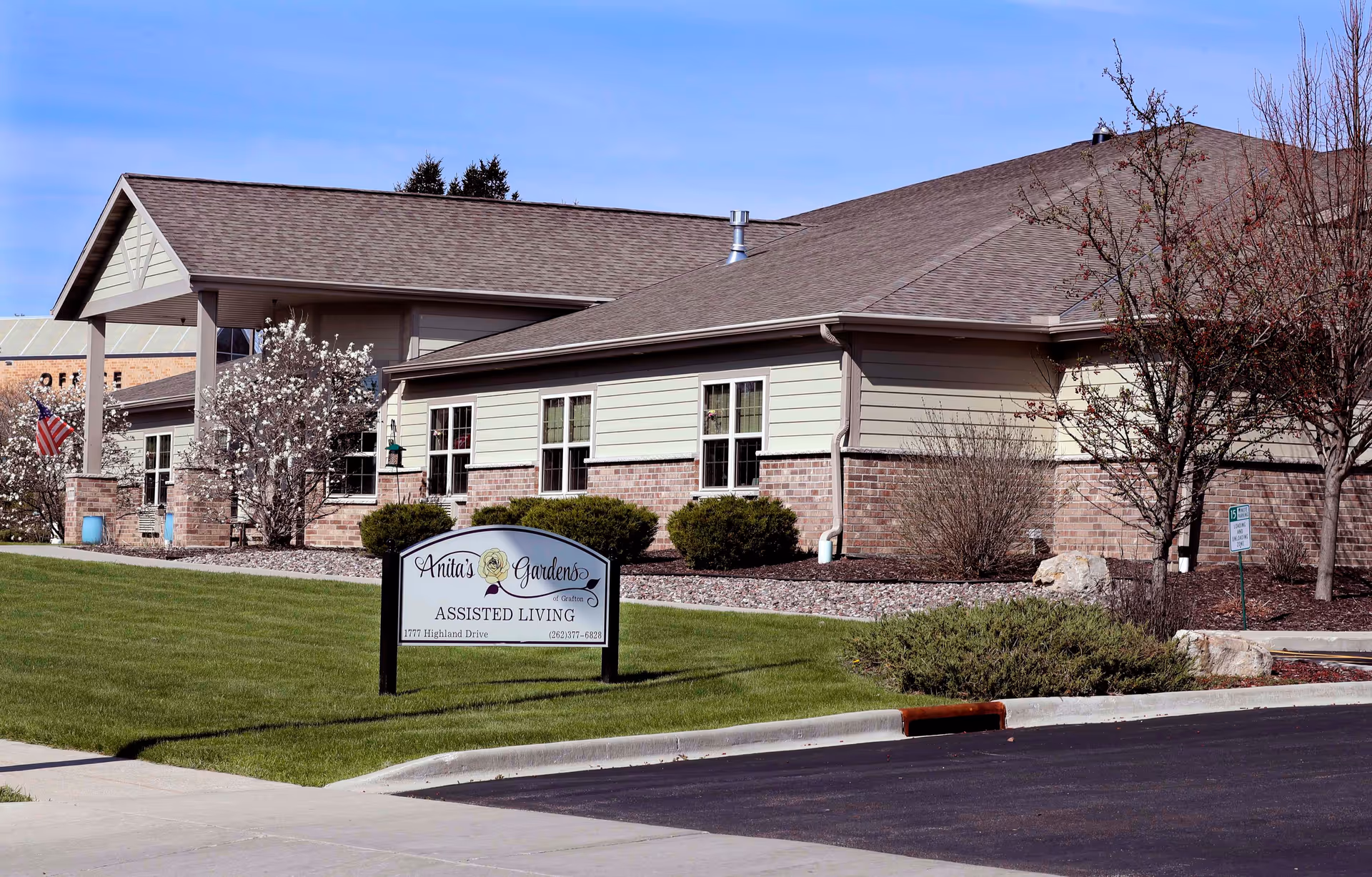 Exterior view of Anita's Gardens of Grafton assisted living facility showing a single-story building with beige siding and brick accents, surrounded by landscaped bushes and trees. A sign in front displays the facility name, address, and phone number. The sky is clear and blue.