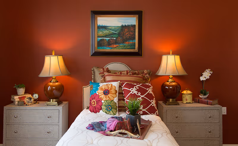 A cozy bedroom with a single bed against a red wall. The bed is decorated with multiple colorful pillows and a tray with a plant and a scarf on the white bedspread. On either side of the bed are matching nightstands with brown lamps, books, and decorative items. Above the bed hangs a framed landscape painting.