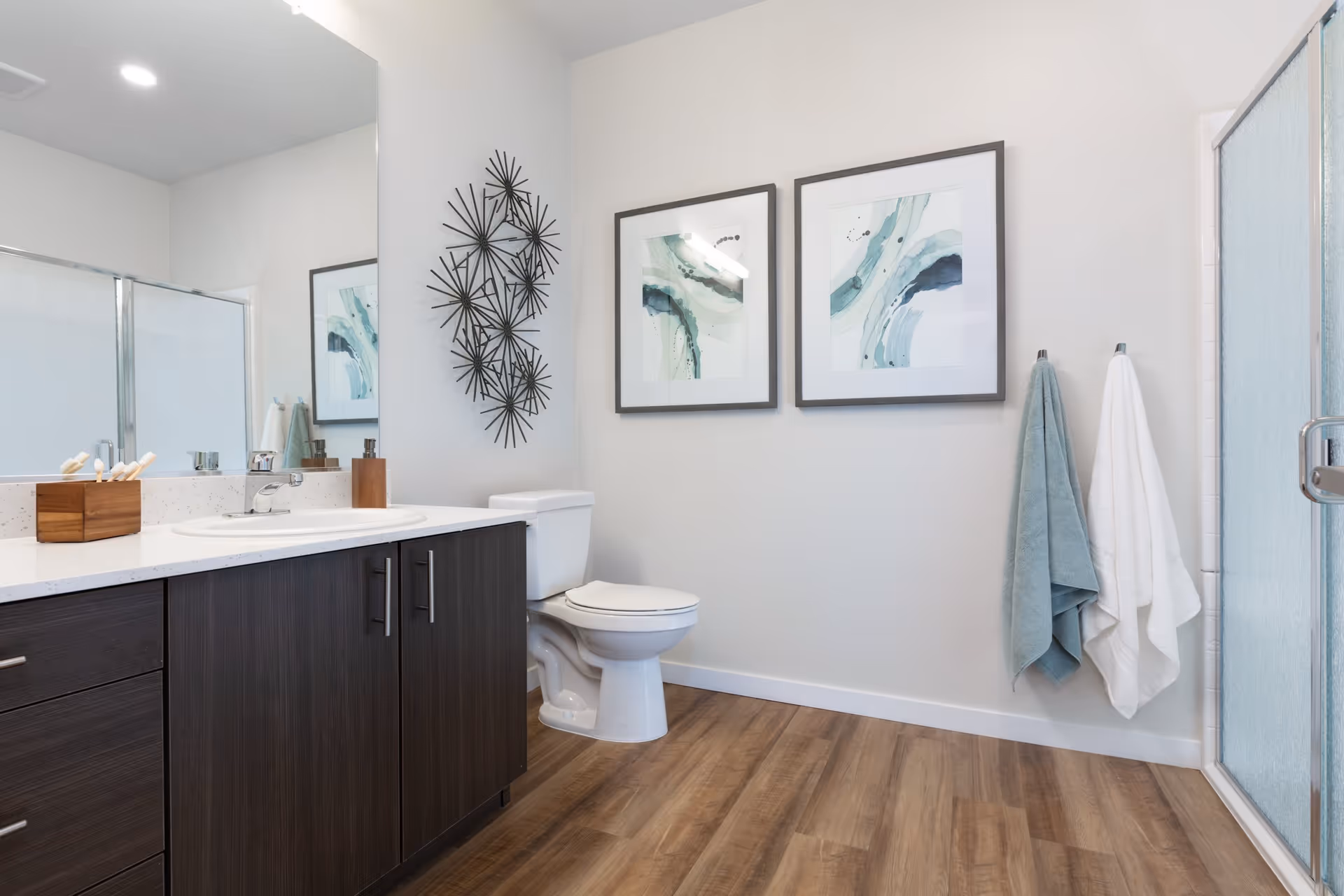A modern bathroom with a wooden floor, dark wood vanity with a white countertop and sink, a toilet, two framed abstract art pieces on the wall, a metal wall decoration, two towels hanging on hooks, and a glass-enclosed shower.