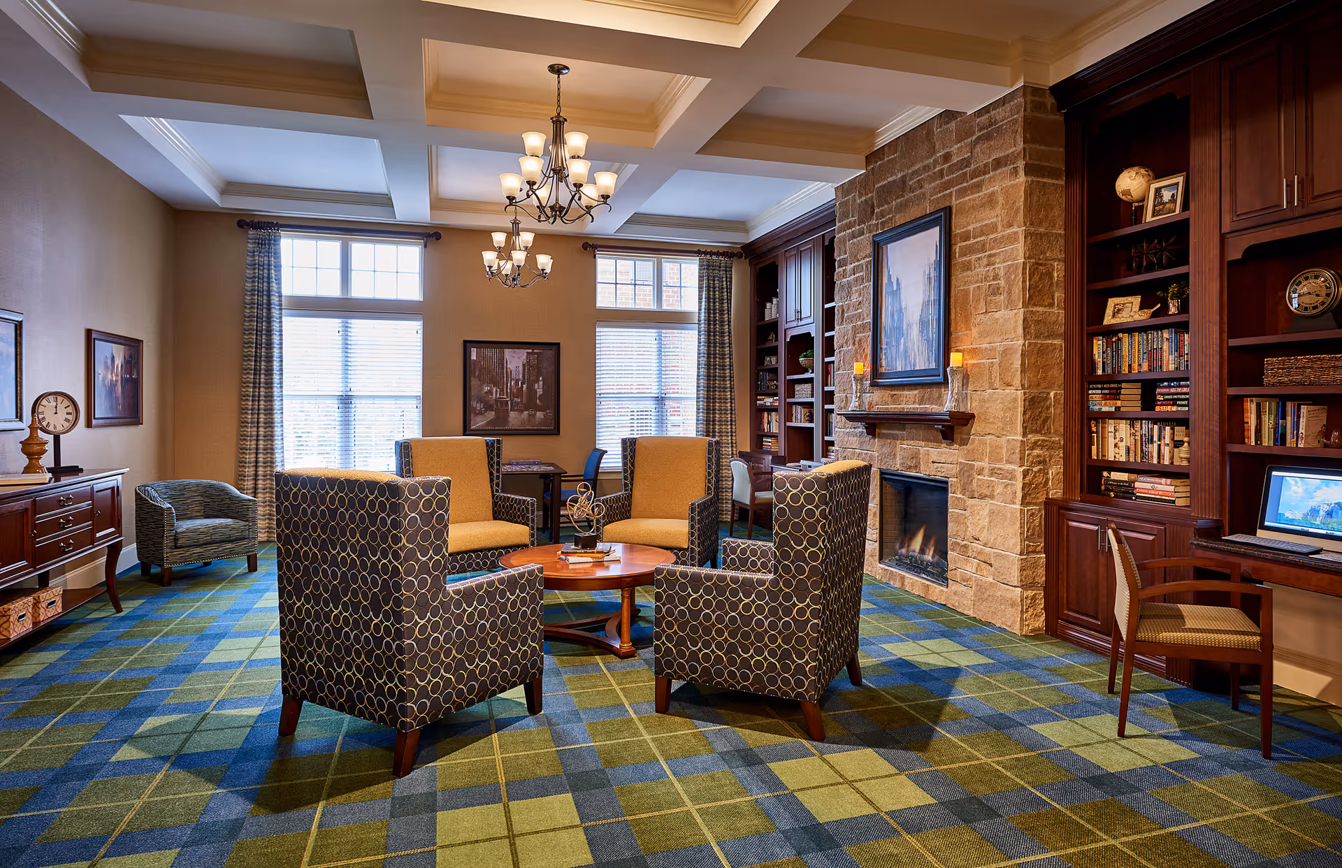 Cozy communal lounge with patterned armchairs arranged around a coffee table, a stone fireplace, built-in bookshelves and large windows.
