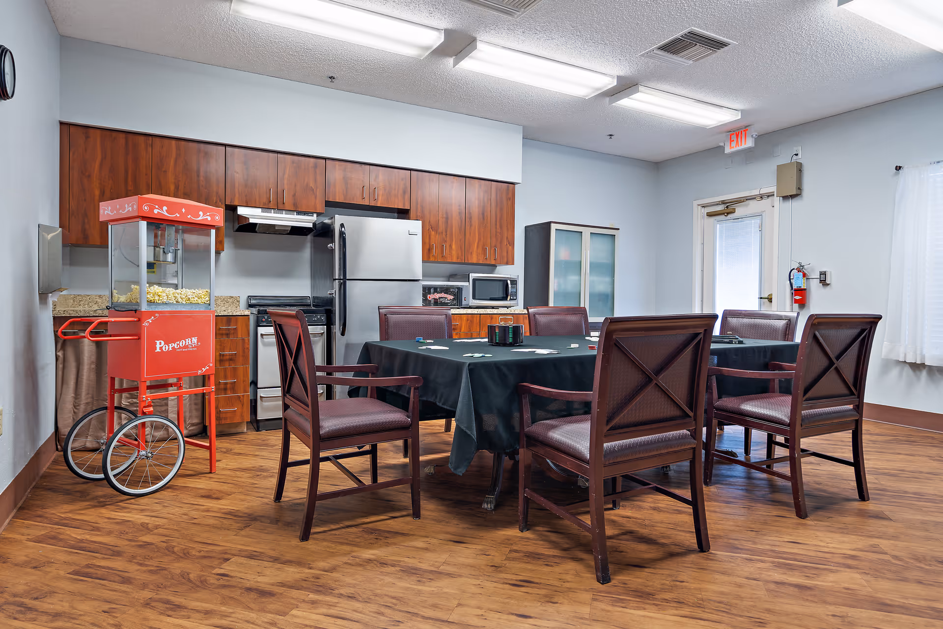 A communal kitchen and dining area with wooden cabinets, a stainless steel refrigerator, stove, and microwave. There is a red popcorn machine on wheels to the left, and a table covered with a black tablecloth surrounded by six wooden chairs with padded seats. The room has wood flooring, white walls, and fluorescent ceiling lights. A door with a window and an exit sign is visible in the background.