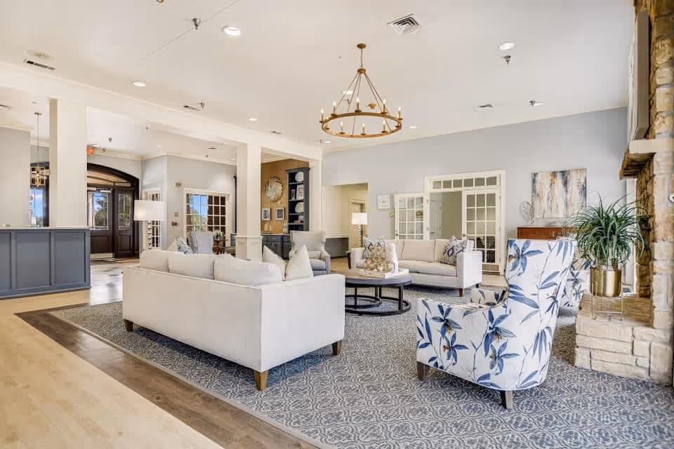 A spacious and well-lit senior living facility common area featuring two white sofas, a round coffee table, a floral-patterned armchair, a stone fireplace, and decorative plants. The room has light-colored walls, a patterned carpet, and a chandelier hanging from the ceiling.